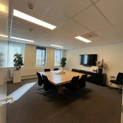 Modern ingerichte vergaderruimte in een kantoorpand aan de Catharina van Renneslaan 8-10, Hilversum, ideaal om te huren. Ruime tafel omringd door comfortabele stoelen, met ramen die natuurlijk licht binnenlaten.