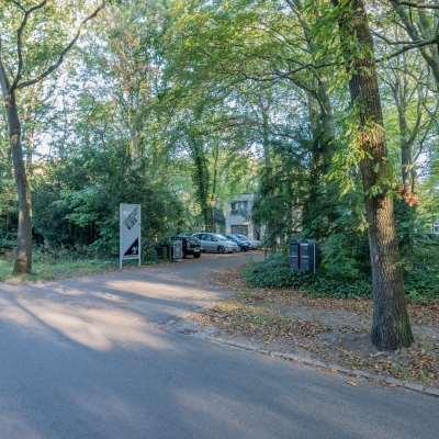 Buitenzicht op het kantoorpand gelegen aan de groene Mozartlaan 25 in Hilversum, omgeven door bomen en met een parkeerplaats vooraan.