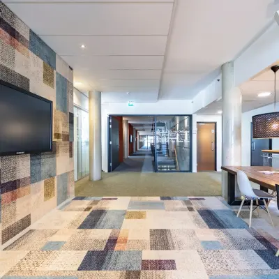 Moderne kantoorruimte te huur op Opaallaan 1180 in Hoofddorp met kleurrijk tapijt en grote wandtelevisie, voorzien van houten tafel en stoelen.