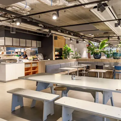 Moderne kantoorruimte in Hoofddorp met gemeubileerde werkplekken, een open keuken en groene accenten. Mensen zien bezig met het bereiden van eten. Ideaal om een kantoorruimte te huren.