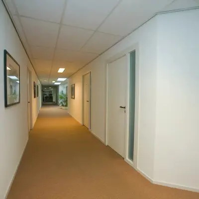Verlichte gang met bruin tapijt en witte muren in kantoorpand op Sandtlaan 36, Katwijk. Verschillende deuren zichtbaar. Kantoorruimte huren mogelijk.