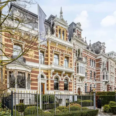 Rij monumentale 19e-eeuwse herenhuizen in neorenaissancestijl met gestreepte rode en gele bakstenen gevels, torentjes en sierlijke balkons, achter een zwart smeedijzeren hek met buxushagen, met moderne hoogbouw op de achtergrond.