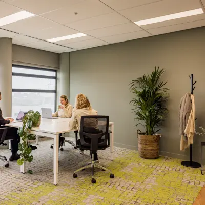 In deze moderne kantoorruimte op Wilhelminaplein 1-40, Rotterdam Kop van Zuid, werken drie personen met laptops aan een witte tafel. De ruimte is stijlvol ingericht met planten en grote ramen.