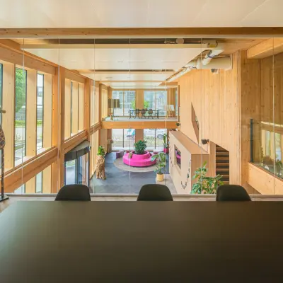 Moderne kantoorruimte met houten interieur en grote ramen, geeft uitzicht op een open zicht van meerdere verdiepingen met kleurrijke meubels en planten, geschikt voor diverse werkactiviteiten.