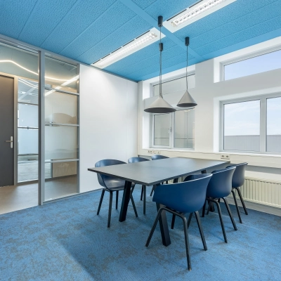 Moderne, goed verlichte kantoorruimte met blauwe accenten, voorzien van een tafel en zes stoelen, ideaal voor vergaderingen of als werkruimte. Grote ramen bieden veel natuurlijk lichtinzichten. Geschikt voor kantoorpand huren.